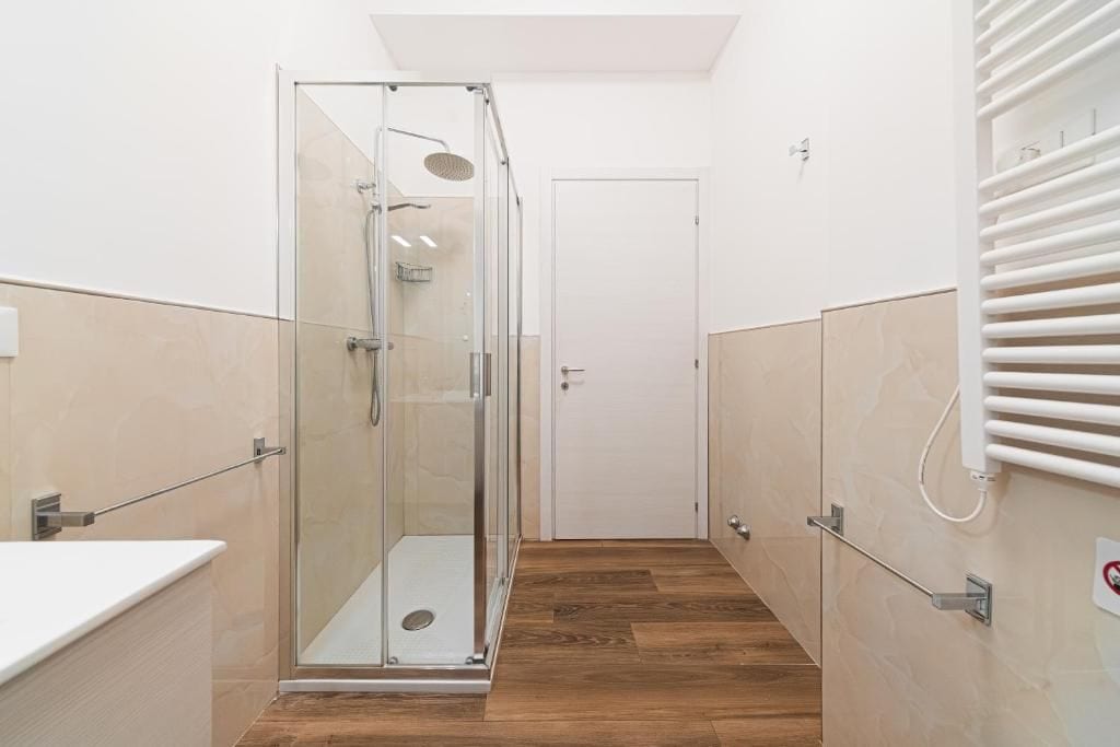 A narrow hallway with a glass-enclosed shower area, a white door at the end, and a towel rack on the right wall.