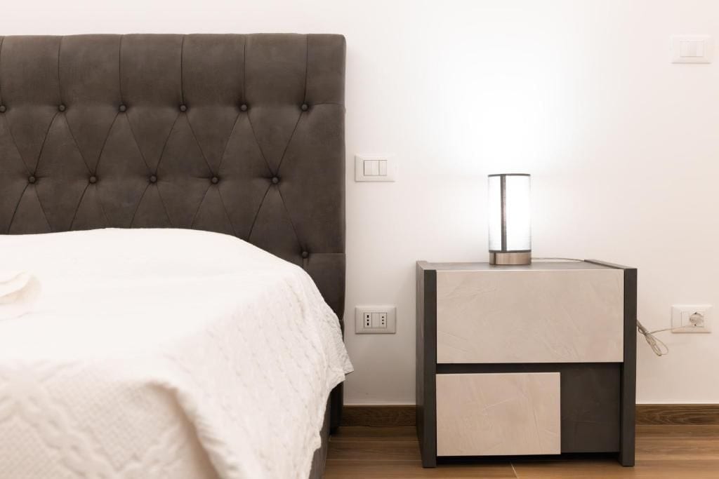 Ein Bett mit einem gesteppten Kopfteil, ein Nachttisch mit einer Lampe und eine Steckdose an der Wand.