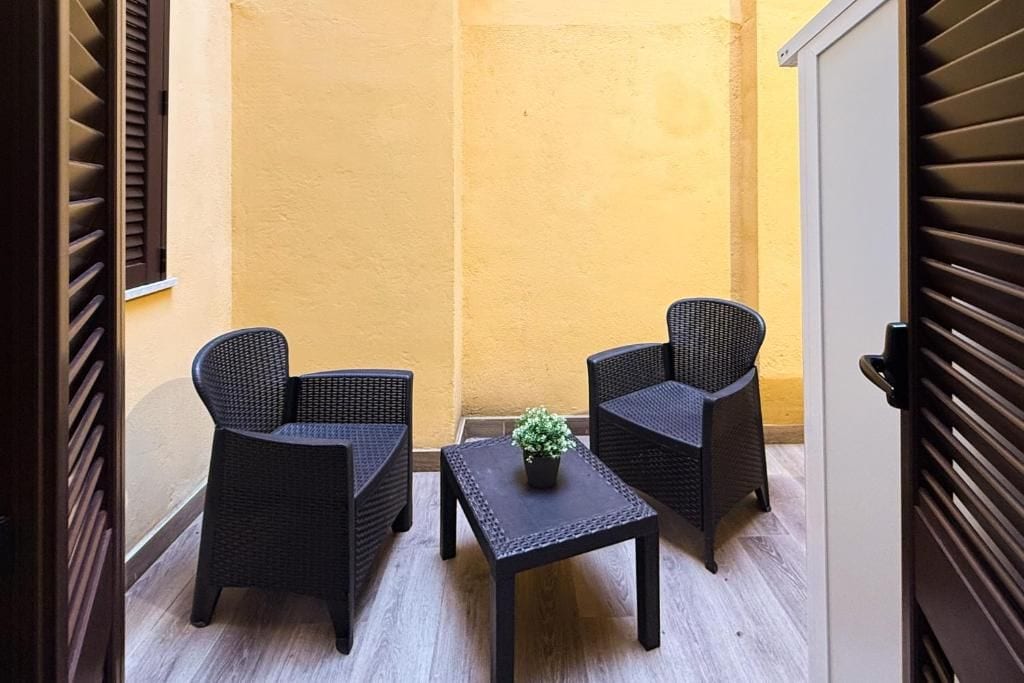 Un pequeño espacio de estar al aire libre con dos sillas de mimbre y una mesa, una planta en maceta sobre la mesa, y una pared amarilla al fondo.