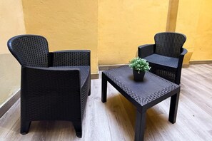 Uma pequena área externa com assentos, duas cadeiras de vime e uma mesa, um vaso com planta na mesa, e uma parede amarela ao fundo.