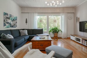 A modern living room with a dark blue sofa, a wooden coffee table, and a grey ottoman. There is a large window with curtains, a potted plant, and a wall-mounted television. - Aurora Forest House, Sauna By Lapland Host (Rovaniemi)