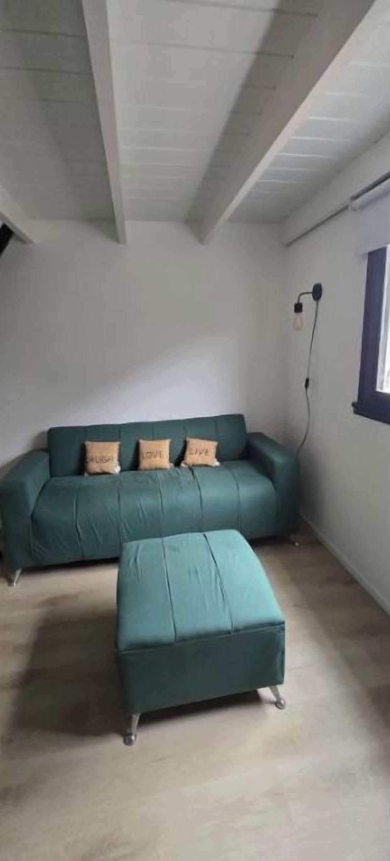 A room with a green sofa and matching ottoman, a window with a view, and a white ceiling with exposed beams.