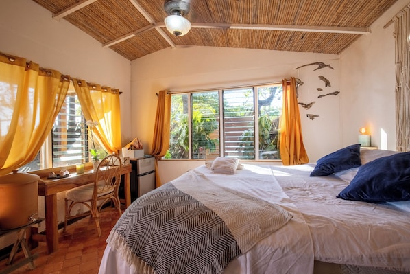 A room with a bed, a desk, and a chair. There is a window with curtains, a ceiling fan, and a wall with a light fixture.
