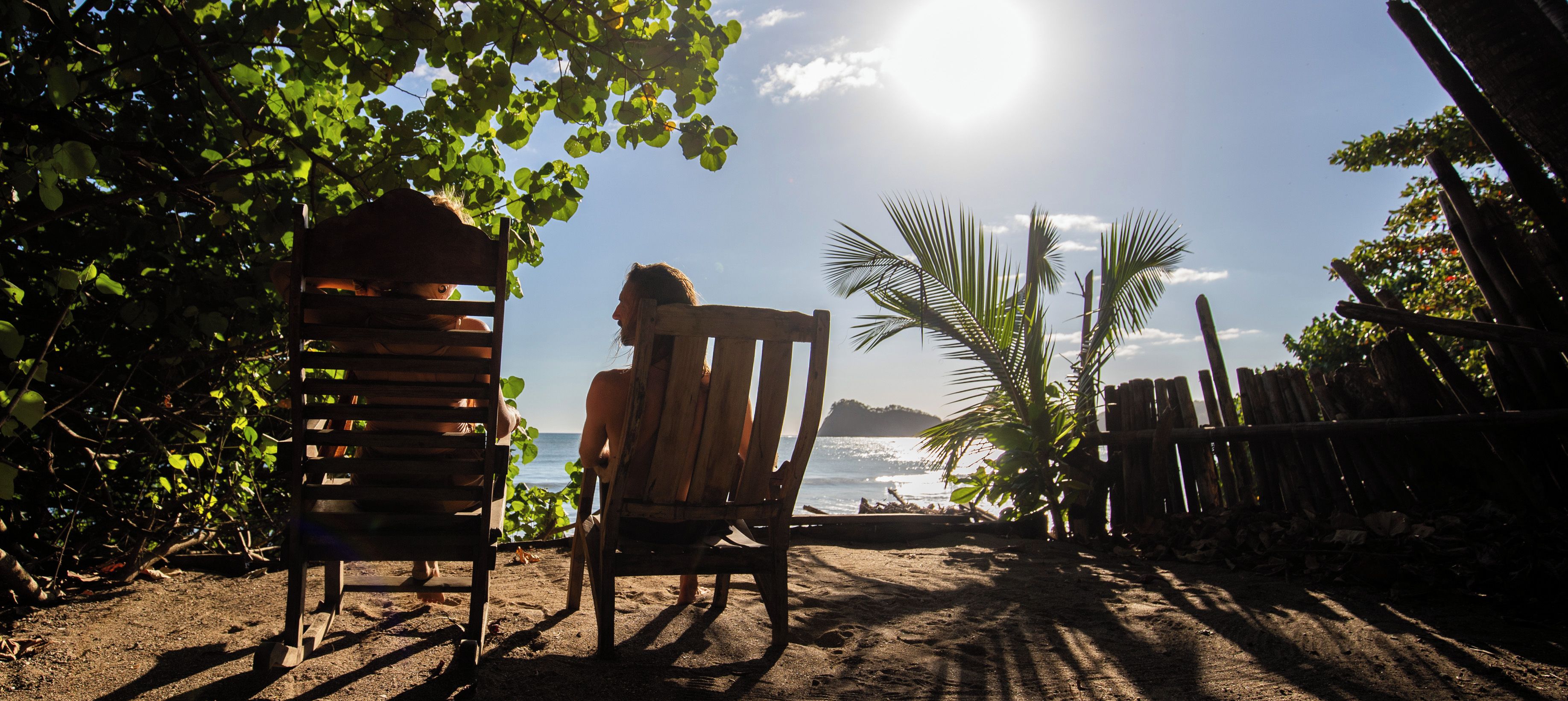Une scène de plage avec une chaise en bois et une structure en bois, peut-être une cabane, avec vue sur la mer et des palmiers.