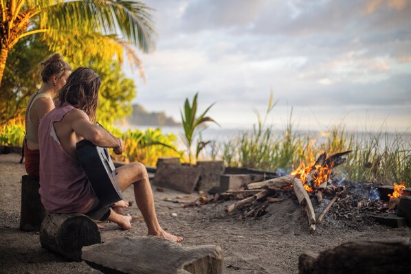 Two people sitting on wooden logs by a campfire with a beach and palm trees in the background.