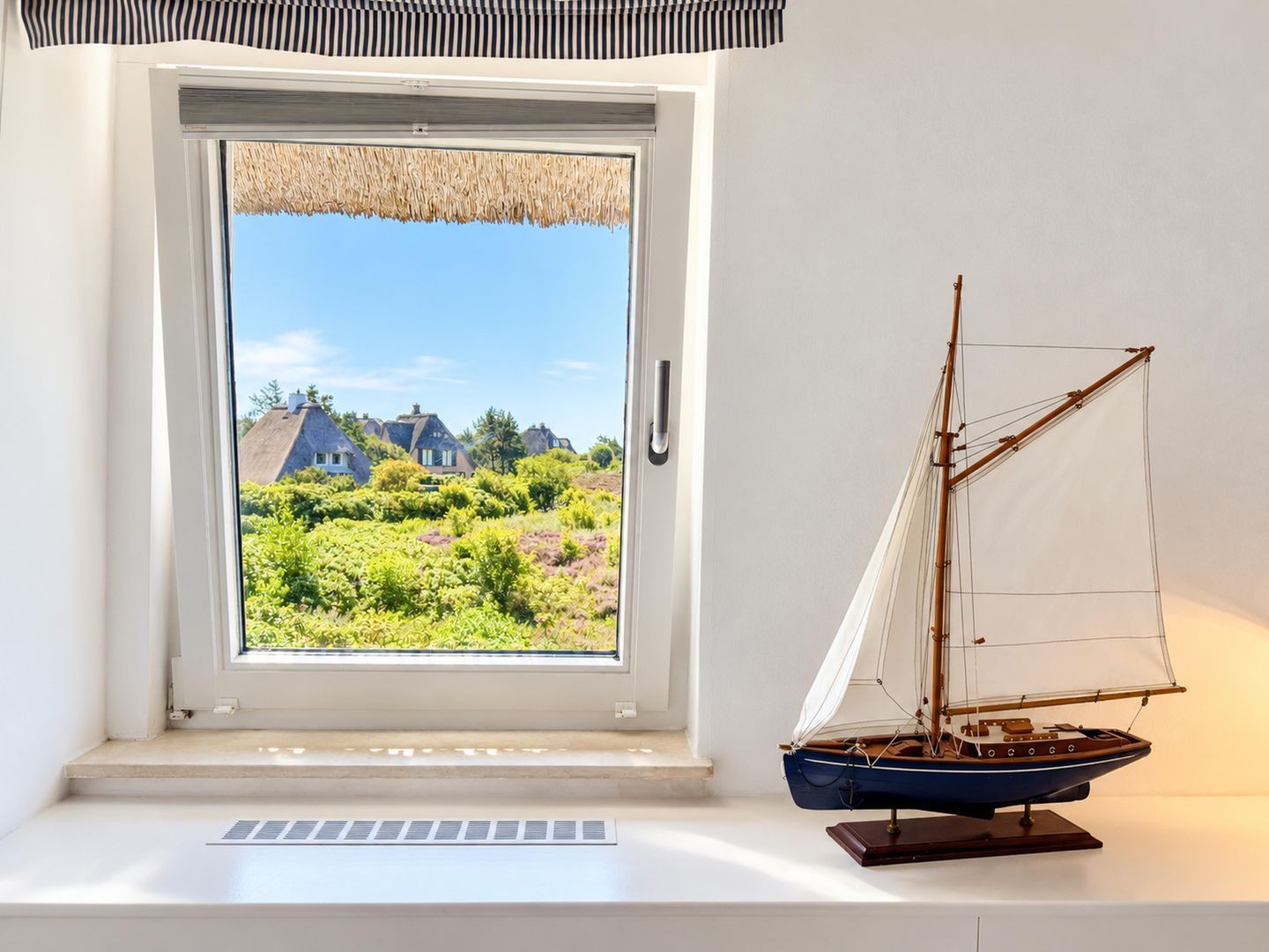 Ventana con vistas a casas y follaje, un velero modelo en un estante y un radiador blanco.