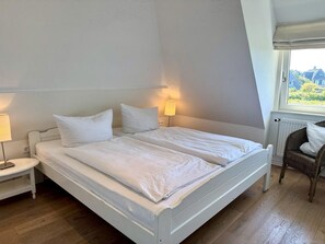 A bedroom with a white bed, two bedside lamps, a wicker chair, and a window with a view. - Haus Westerheide - Vacation home for up to 4 people with a stunning view of the heath (List auf Sylt)