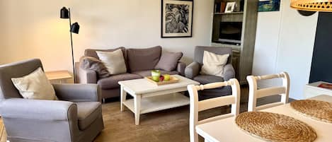 Ein Wohnzimmer mit einer Couch, Sesseln, einem Couchtisch und einem Esstisch mit Platzdeckchen.