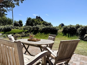 An outdoor seating area with a round wooden table and chairs, surrounded by greenery and a clear sky. - Haus Westerheide - Vacation home for up to 4 people with a stunning view of the heath (List auf Sylt)