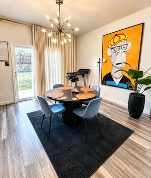 A dining area with a round table, chairs, a black rug, a potted plant, and a large abstract painting on the wall. - Luxury 5BR Designer Home • Sleeps 15 • Near Dallas • Extended & Short Stays (Glenn Heights)