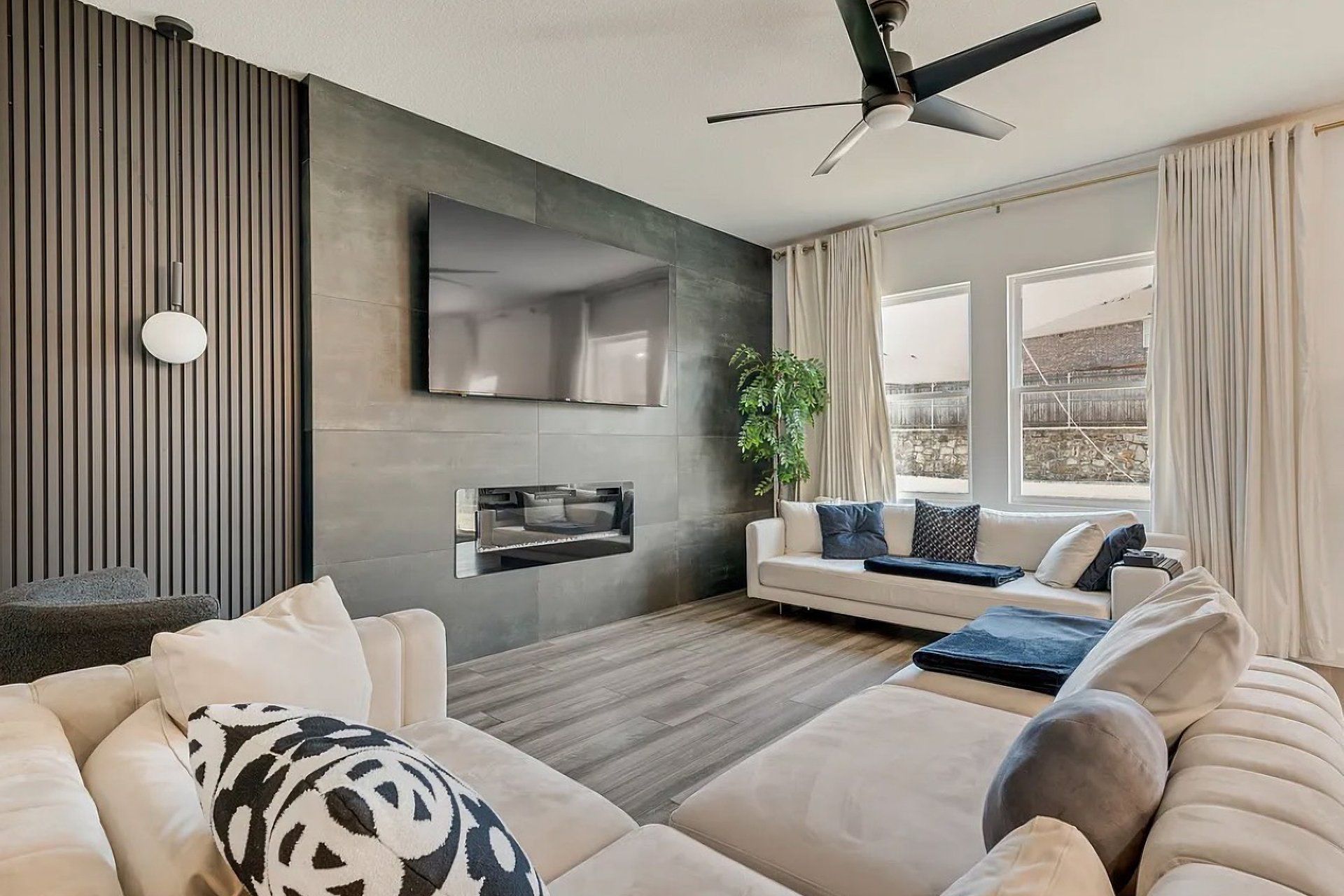 Una sala moderna con sofá, televisor de pantalla plana montado en la pared, ventilador de techo y un ventanal con cortinas.