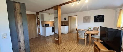 Un espace de vie moderne avec un plancher en bois, un coin repas comprenant une table et des chaises, un téléviseur et une cuisine en arrière-plan.
