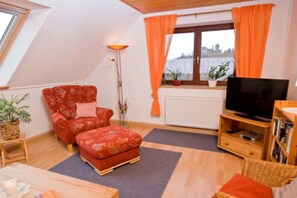 A cozy living room with a red armchair, a matching ottoman, a television on a wooden stand, and a view of the city through the window. - Holiday apartment with 3 bedrooms (Colmberg)