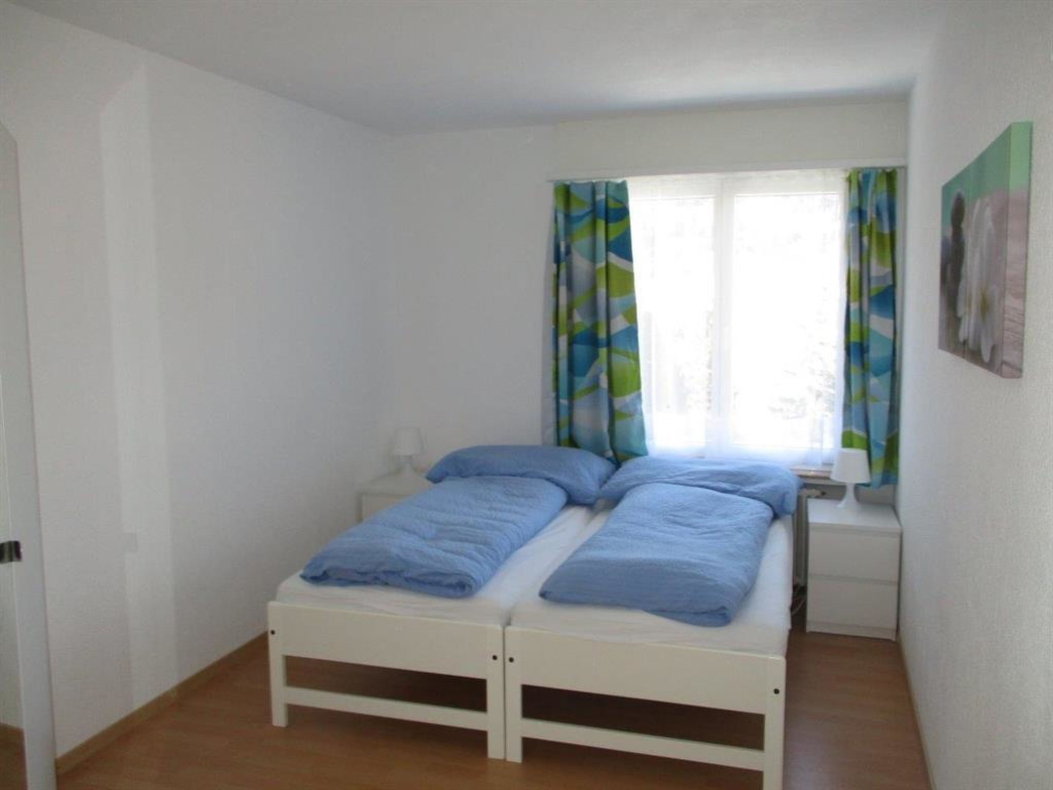 A small, single-bed room with a window and curtains, a bedside table, and a wall-mounted picture.