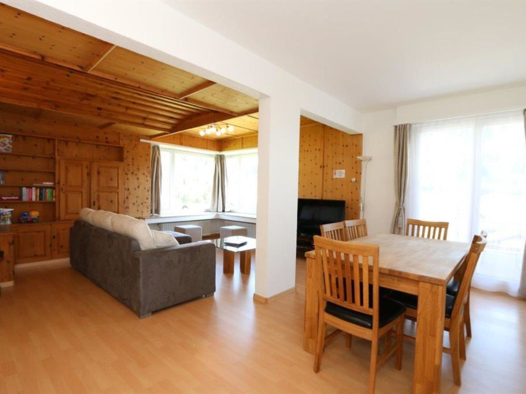 Ett rymligt vardagsrum med soffa, matbord och stolar, träpanel på väggarna och ett köksområde.