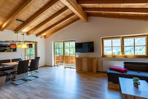 A spacious living area with a high ceiling, wooden beams, a dining table with chairs, a sofa, a TV mounted on the wall, and large windows with a view of greenery. - Holiday home with 1 bedroom (Marktschellenberg bei Berchtesgaden)