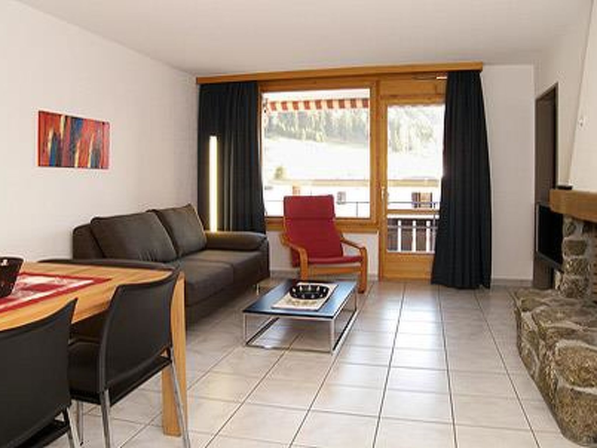 Ein Wohnzimmer mit einer Couch, einem Esstisch mit Stühlen, einem roten Sessel und Blick durch eine Glastür nach draußen.