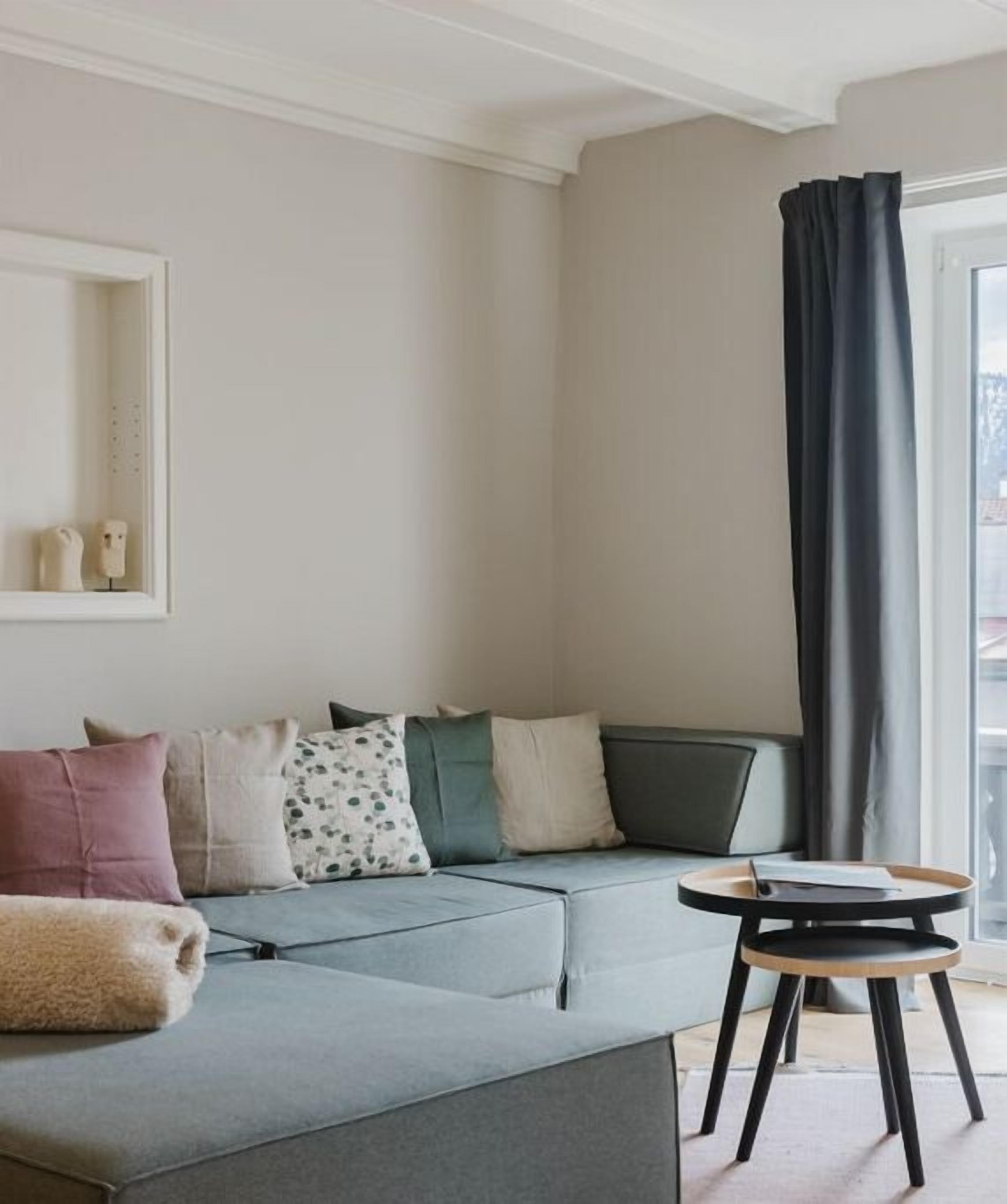 Un salon moderne avec un canapé d’angle, une petite table ronde et une grande fenêtre agrémentée de rideaux.