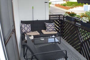 A balcony with a black metal railing, a black and white patterned sofa with cushions, and potted plants with flowers. - Holiday apartment with 1 bedroom (Selb-Plößberg)
