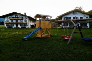 Un'area giochi con uno scivolo e altalene, sullo sfondo di due grandi case con balconi.