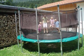 Ein Trampolin mit Sicherheitsnetz, umgeben von einer Rasenfläche und einem Holzstapel.