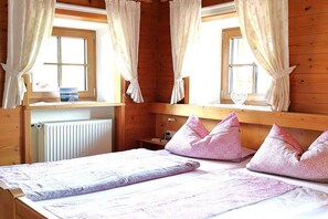 A wooden cabin bedroom with a bed, pillows, a nightstand, a window with curtains, and a radiator. - Apartment with 0 bedrooms (Marquartstein)