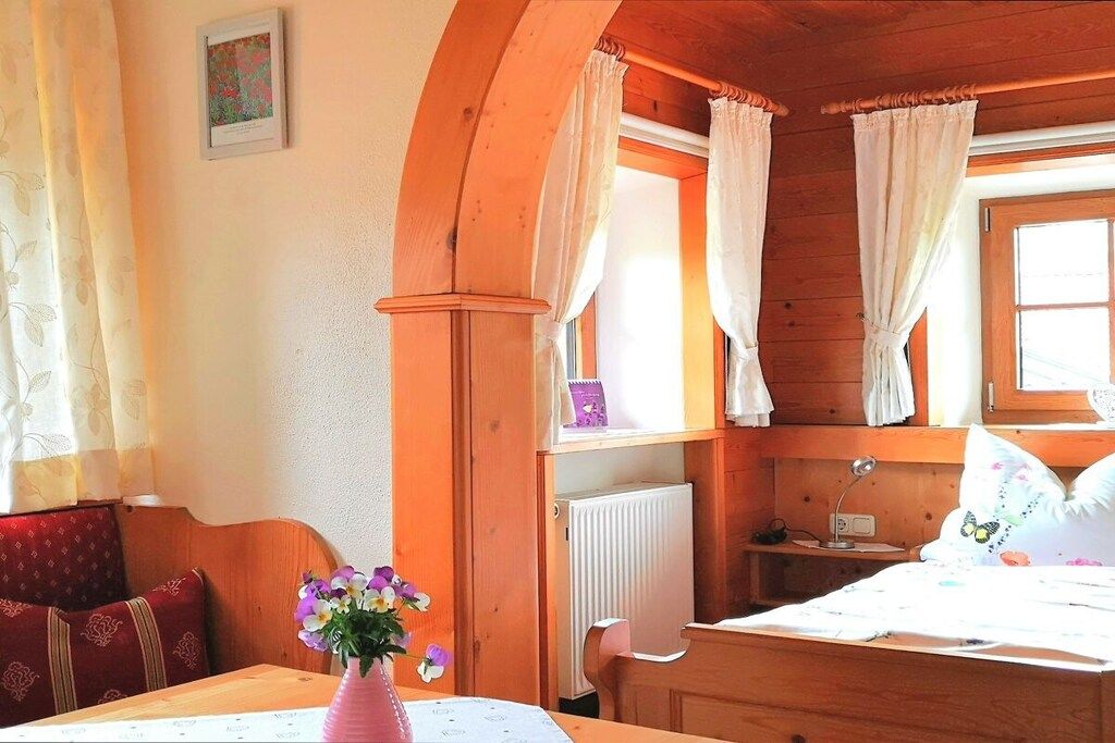 Habitació acollidora amb sostre de fusta, llit amb roba de llit floral, taula amb un ram de flors i finestra amb cortines.