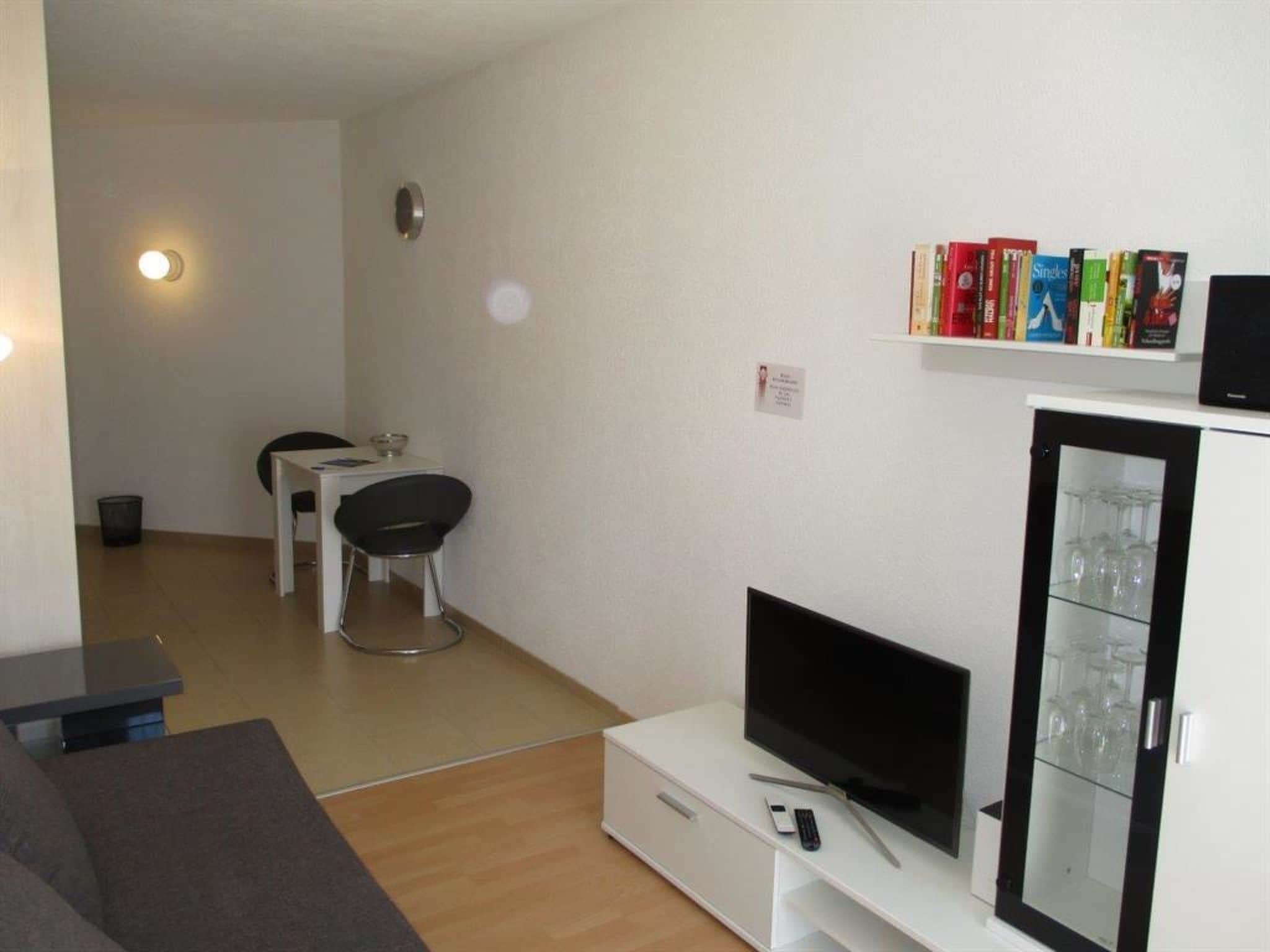 Ein Wohnzimmer mit Flachbildfernseher, einer Bücherwand voller Bücher, einem Essbereich mit Tisch und Stühlen sowie einer Vitrine mit Gläsern.