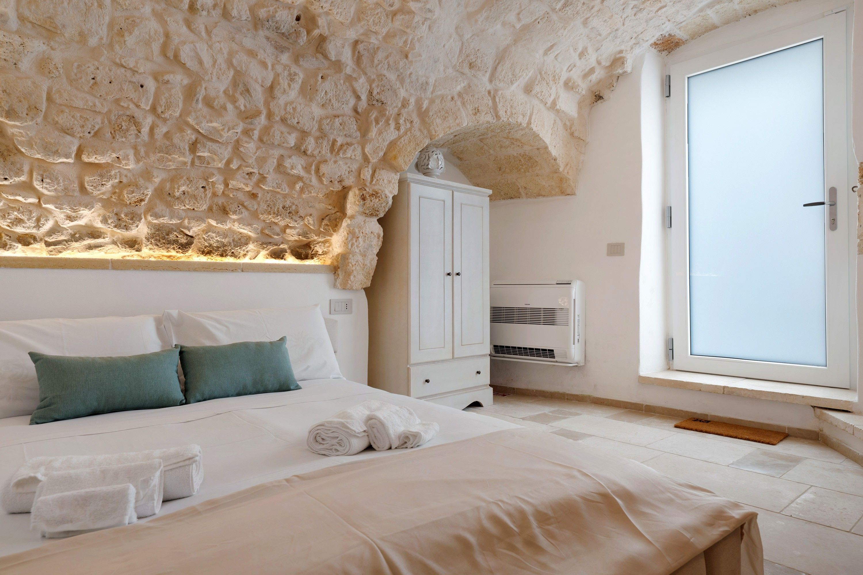 Un dormitorio con pared de piedra, cama con ropa blanca, armario y ventana con vistas.