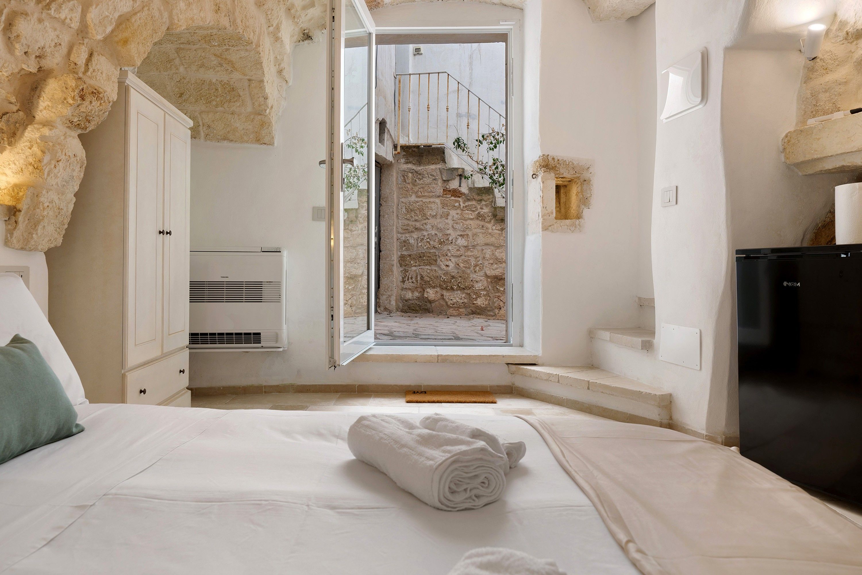 Une chambre avec un lit, une serviette blanche, une armoire, un appareil de climatisation et une vue sur un mur en pierre et un escalier.