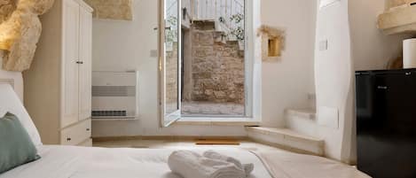 A bedroom with a bed, a white towel, a wardrobe, an air conditioning unit, and a view of a stone wall and staircase.