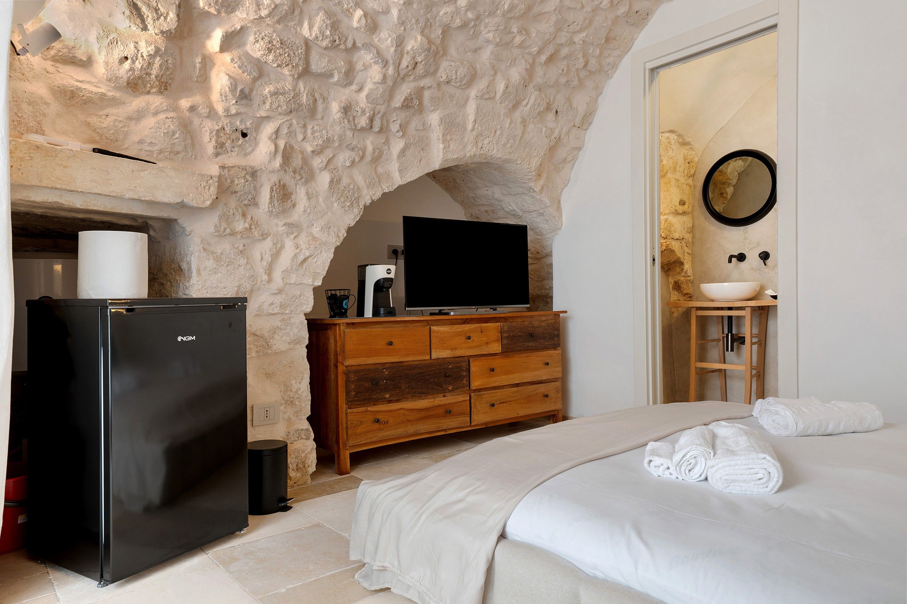 A bedroom with a stone wall, a black refrigerator, a wooden dresser, a television, a sink, and a bed with white linens.