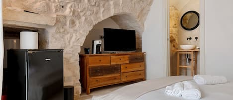 A bedroom with a stone wall, a black refrigerator, a wooden dresser, a television, a sink, and a bed with white linens.