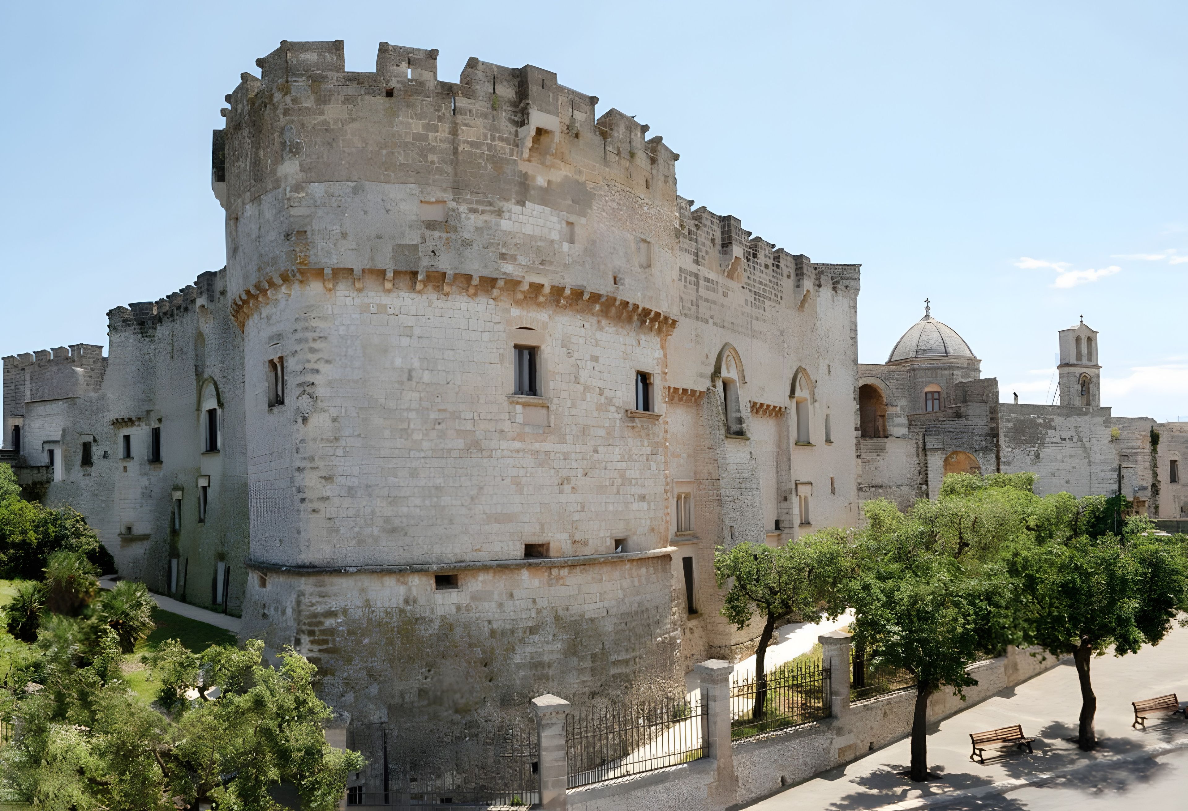 A historic stone castle with battlements, a church with a dome, and a paved walkway with benches.