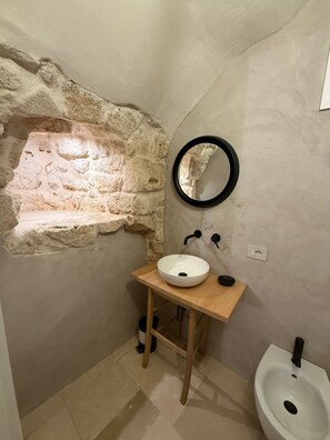 A bathroom with a stone wall, a wooden vanity, a white sink, a black circular mirror, and a white pedestal bidet. - Terra Mia Double Suite Room with Wi-Fi and Air Conditioning (Carovigno)