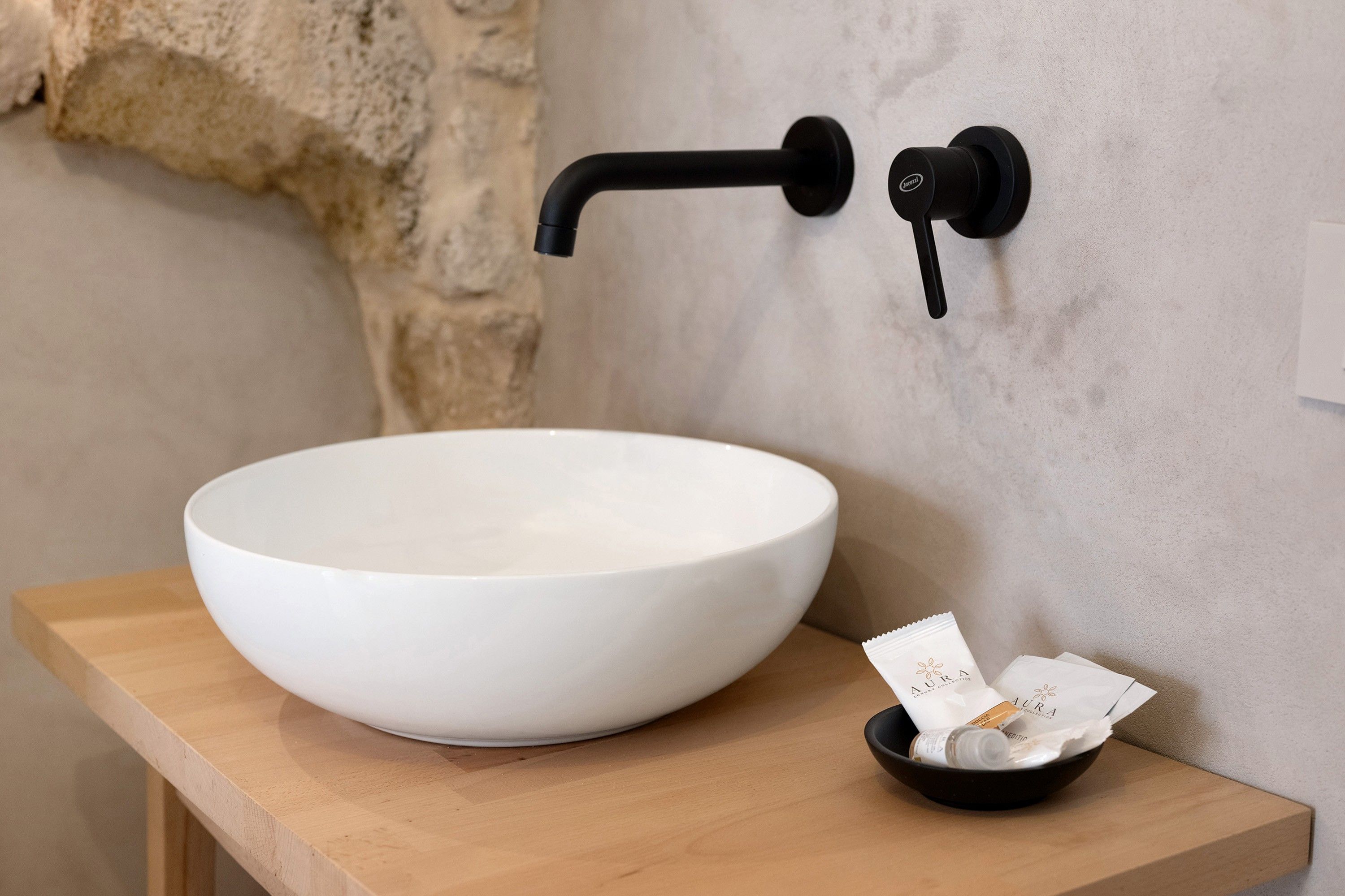 Un lavabo en céramique blanche, posé sur un plan de travail en bois, avec un robinet noir et un petit bol noir contenant un mouchoir et une bouteille.