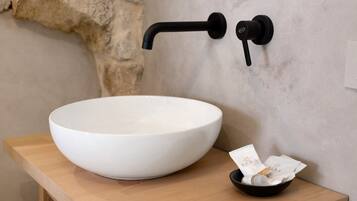 Un lavabo en céramique blanche, posé sur un plan de travail en bois, avec un robinet noir et un petit bol noir contenant un mouchoir et une bouteille.