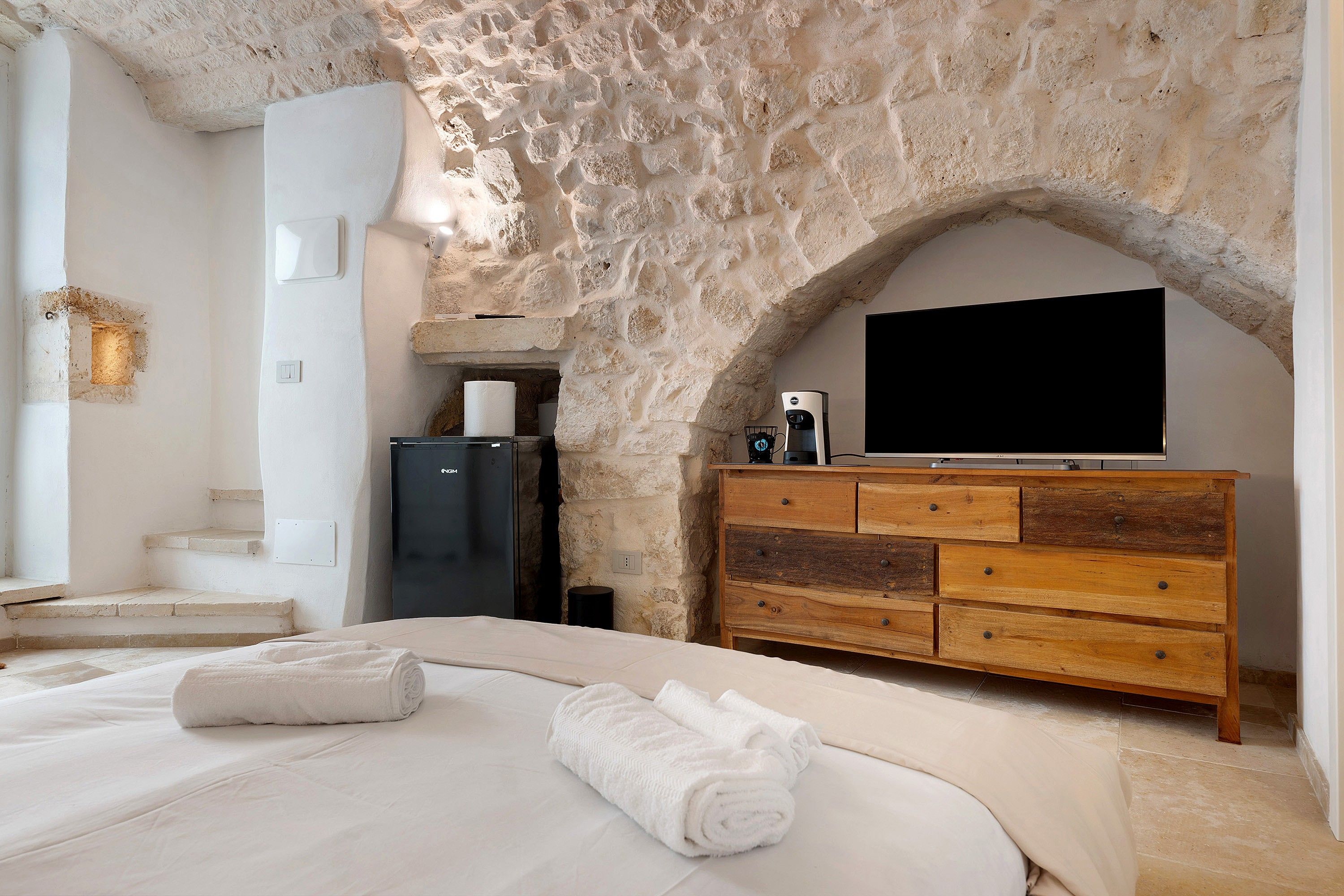 Un dormitorio con pared de piedra, un televisor de pantalla plana, un aparador de madera y una cama con ropa de cama blanca.