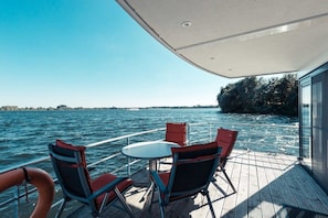 A boat deck with a table and chairs, a railing, and a view of the water and distant shoreline.