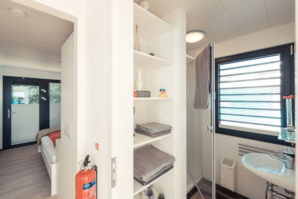 Ein Zimmer mit einem Bett, einem weißen Kleiderschrank mit Regalen, einem Fenster mit Jalousien und einem Badezimmer mit Waschbecken und Spiegel.