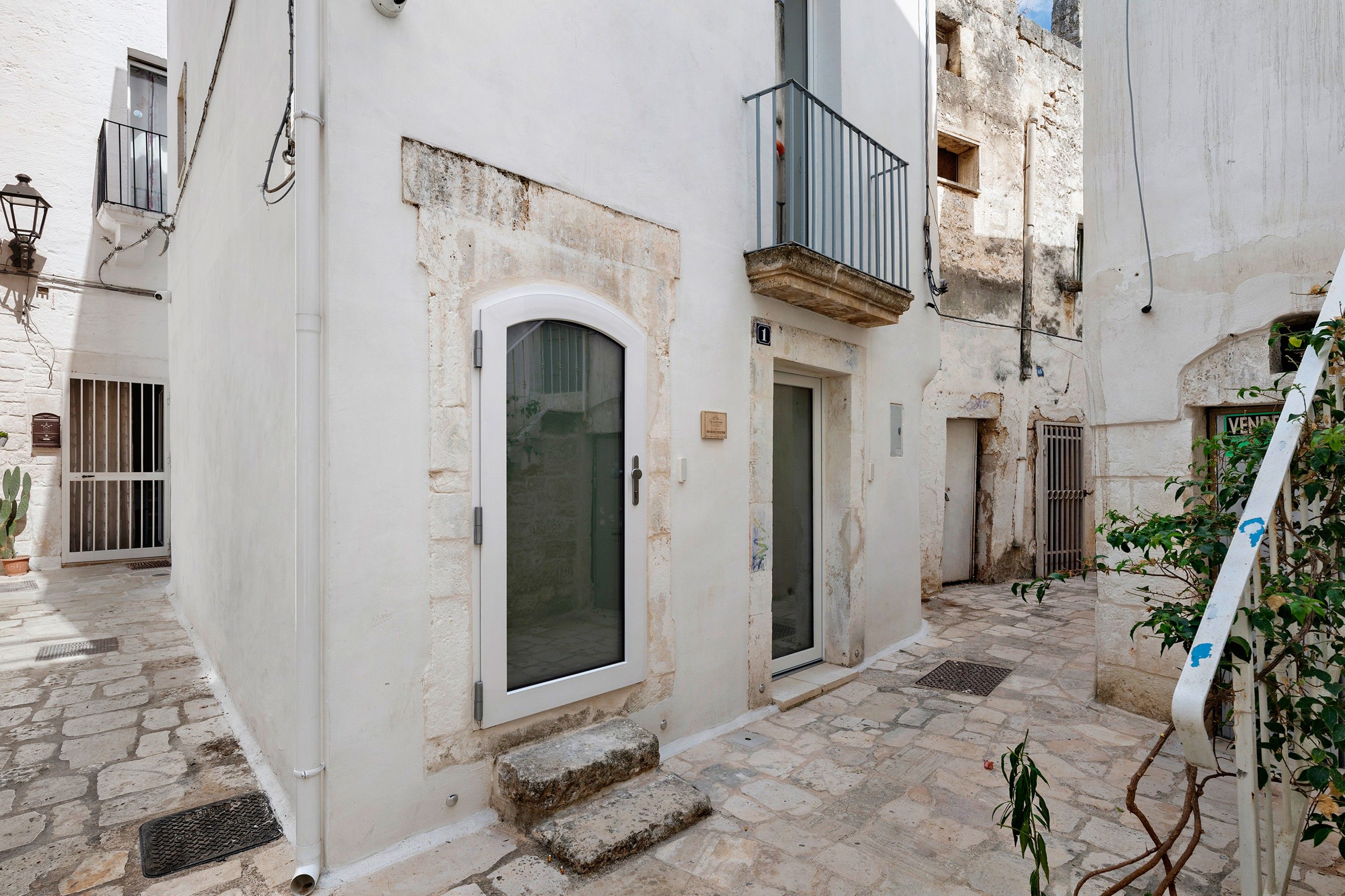 A narrow alleyway with a white building featuring a small door and window, a metal balcony, and stone paving.