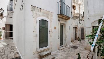 A narrow alleyway with a white building featuring a small door and window, a metal balcony, and stone paving.