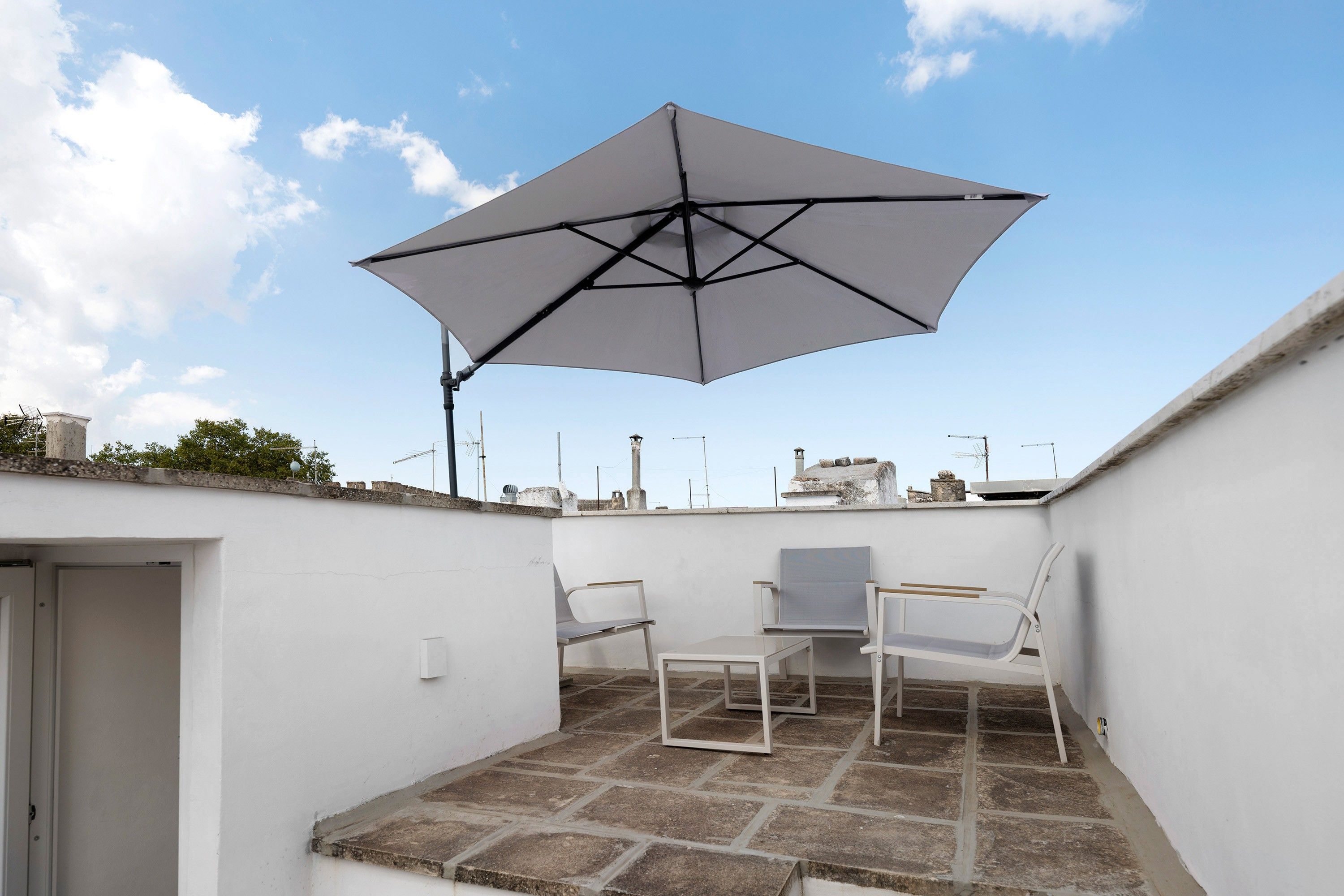 Un patio en la azotea con una sombrilla grande, dos sillas blancas y una mesita.