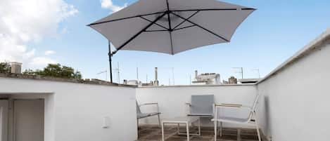 Un patio en la azotea con una sombrilla grande, dos sillas blancas y una mesita.