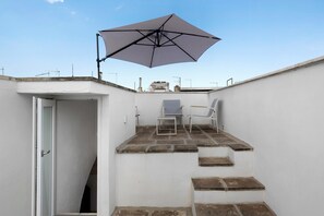 Un pequeño patio al aire libre con una mesa, sillas y un gran paraguas, y una puerta que da a un espacio interior.