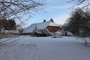 Ein mit Schnee bedecktes Haus mit roter Fassade, umgeben von schneebedeckten Bäumen und Sträuchern.