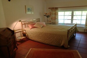 Ein Schlafzimmer mit Bett, Kommode, Nachttisch, einem Fenster mit Vorhängen und einem Bild an der Wand.