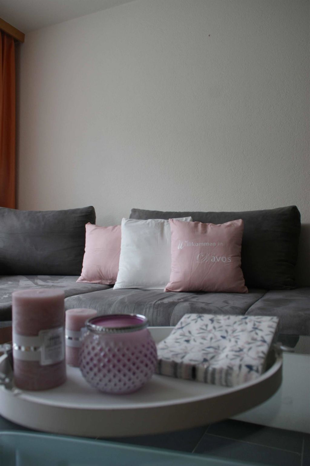 Una sala de estar con un sofá gris, un cojín blanco y rosa, una bandeja con velas y un cojín estampado.