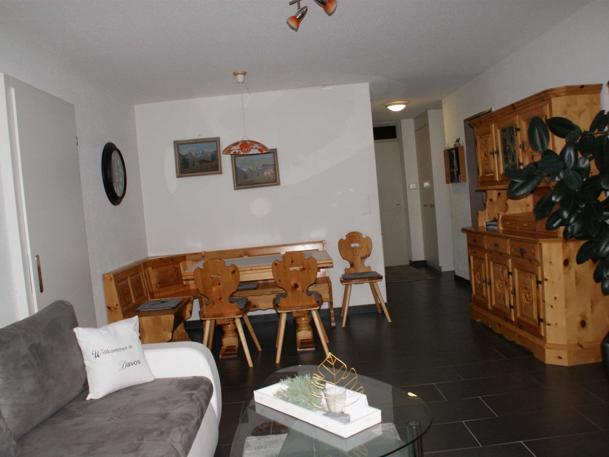 Una sala de estar con un sofá, una mesa de centro, una zona de comedor con sillas de madera y un banco, y una cocina con armarios de madera y una planta.