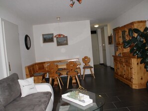 Ein Wohnzimmer mit einer Couch, einem Couchtisch, einem Essbereich mit Holzstühlen und einer Bank sowie einer Küchenzeile mit Holzschränken und einer Pflanze.